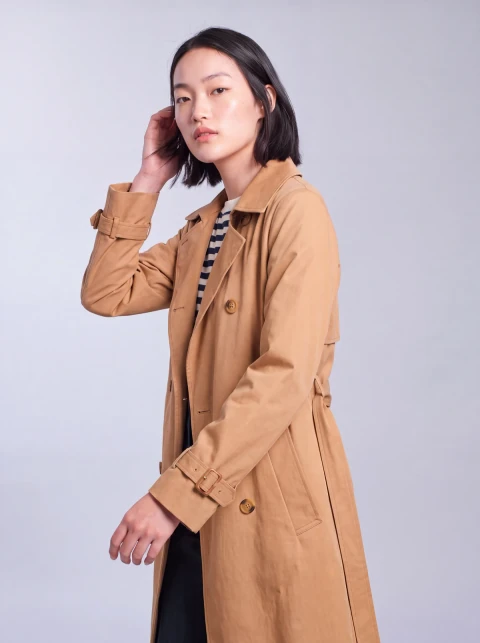 Woman turning in a camel trench coat over a striped top against a pale studio background.